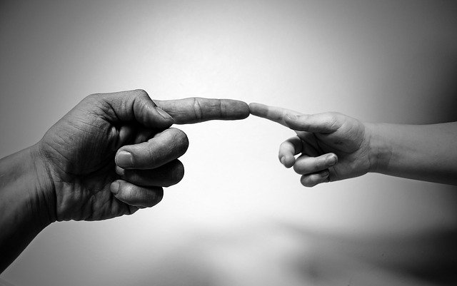 Foto en blanco y negro, muestra la mano de un adulto apuntando, y tocando el dedo de un menor, que también está apuntando.